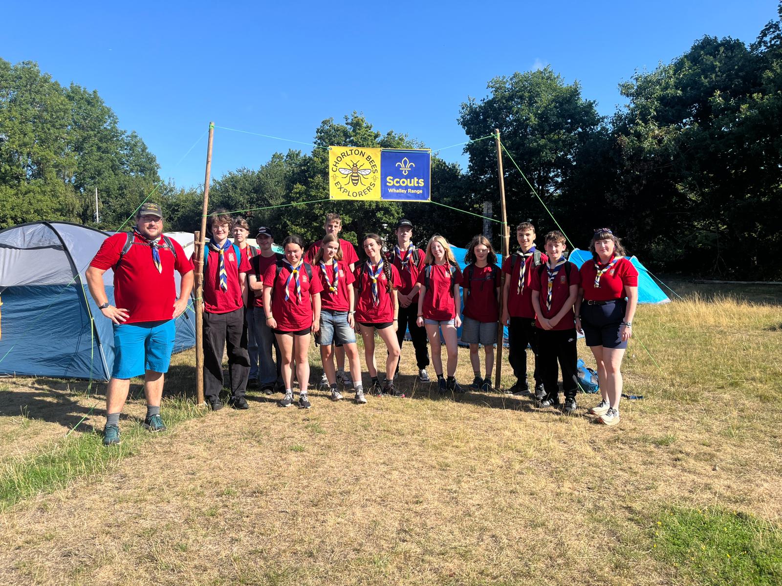 Photograph of group of explorers posting for a photograph in front of a tent and a Scouts banner, with brown grass in the foreground and trees and blue sky in the background